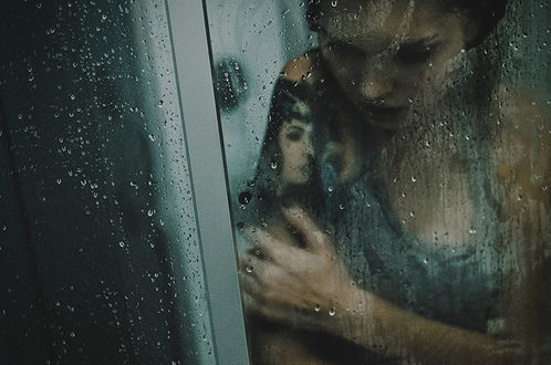 Обои на рабочий стол Темноволосая девушка с татуировкой женского лика на предплечье, стоящая за стеклом с каплями воды