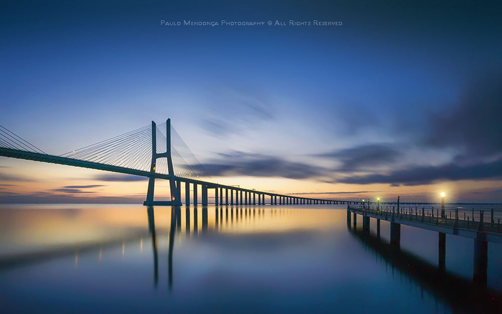 Обои для рабочего стола Автомобильный мост, проходящий через морской залив и каменный пирс, уходящий от берега на фоне желтой полоски заката на вечернем небосклоне с синими облаками, автор PAULO MENDONCA