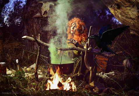 Обои на рабочий стол Рыжеволосая колдунья, стоящая возле костра с кипящим на огне волшебным зельем, рядом на деревянной перекладине сидит черный ворон, вокруг костра разбросаны предметы для колдовства, автор Геннадий Клименко