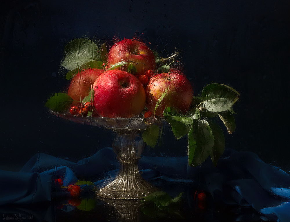 Обои для рабочего стола Ваза с красными, спелыми яблоками и темно-зелеными листьями стоящая на столе с синей драпировкой за оконным стеклом, покрытом дождевыми каплями