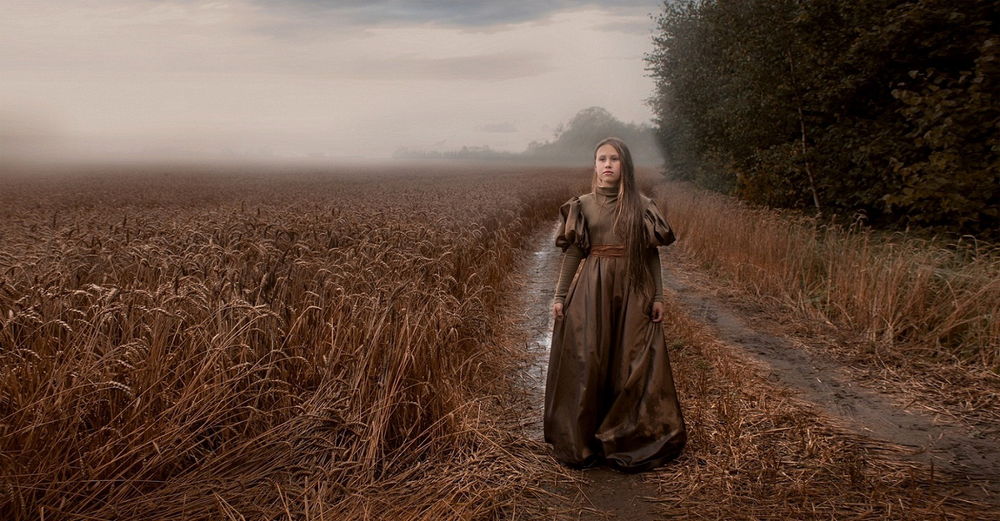 Обои для рабочего стола Девочка в темном, длинном платье, стоящая на дороге, проходящей через поле спелой ржи на фоне пасмурного неба с туманной дымкой