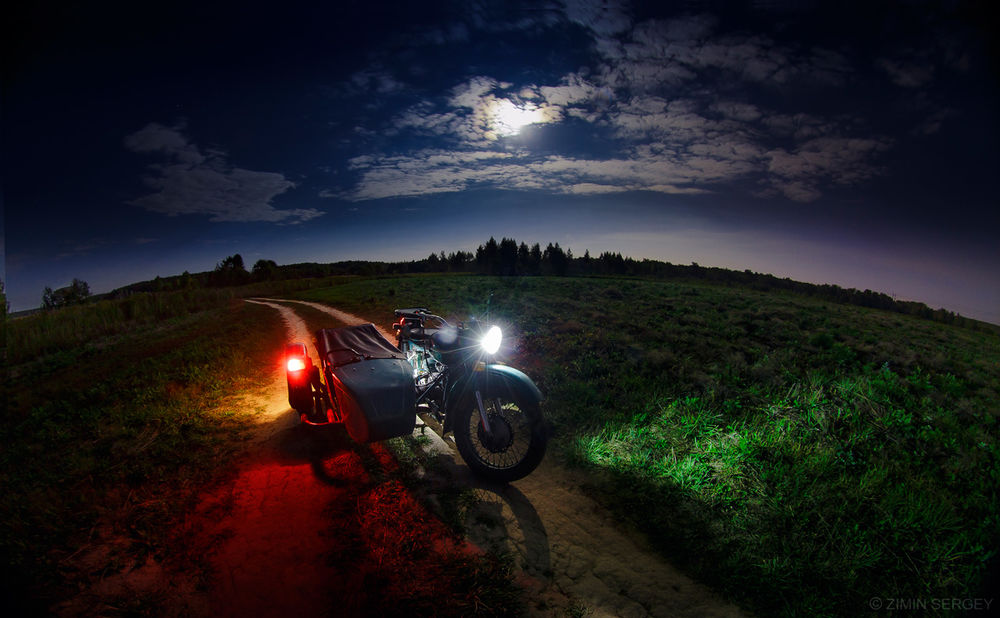 Обои для рабочего стола Мотоцикл, с включенными фарами, стоящий на грунтовой дороге, проходящей по зеленому полю на фоне ночного неба с луной, спрятавшейся за облака, фотография выполнена с помощью широкоугольного объектива фотографом Сергеем Зиминым