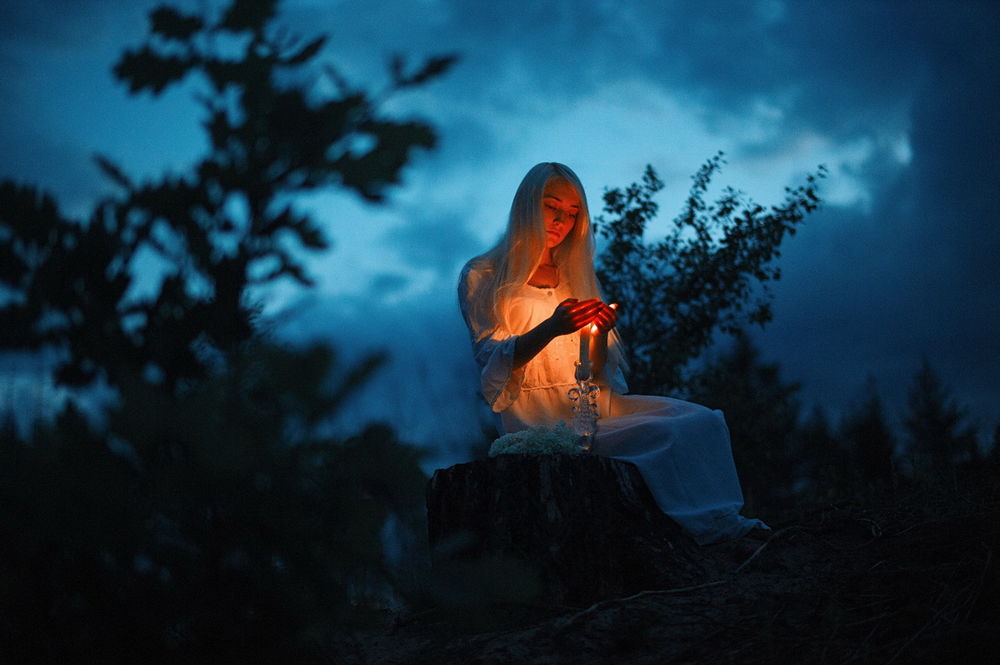 Обои для рабочего стола Светловолосая девушка в длинном, белом платье, сидящая на пеньке, держащая ладони рук над горящей свечой на фоне ночного, пасмурного неба