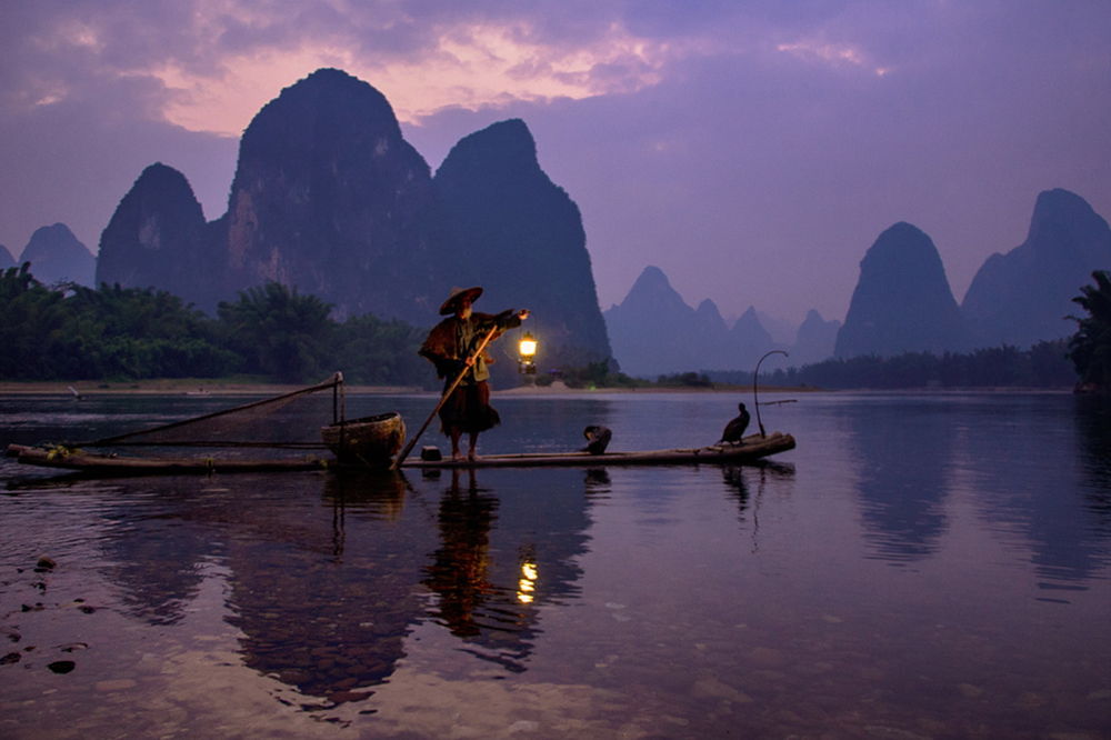 Обои для рабочего стола Пожилой китайский рыбак, стоящий на узком, деревянном плоту, держащий в одной руке деревянный шест, а в другой зажженный фонарь, находящийся на мелководье горного озера на рассвете, на носу плота сидит баклан