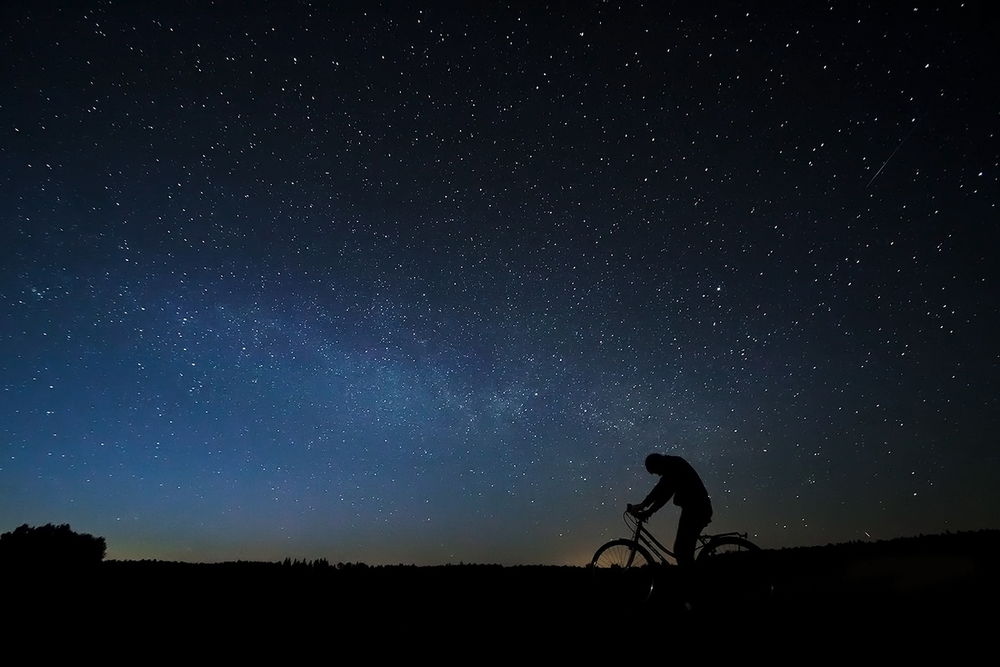 Обои для рабочего стола Мужчина с опущенной головой, сидящий на велосипеде на фоне ночного, звездного неба и Млечного пути