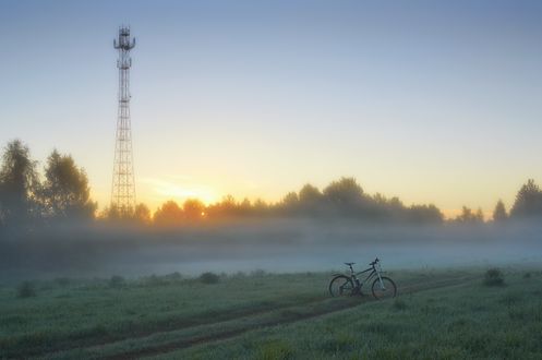Обои на рабочий стол Велосипед, стоящий на дороге, проходящей по зеленому полю с лесопосадками на рассвете на фоне восходящего солнца на безоблачном небосклоне, телевизионной вышки, автор Александр Виноградов
