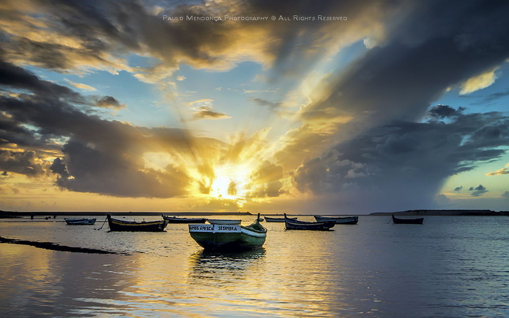 Обои для рабочего стола Прогулочные лодки, стоящие на мелководье морского побережья на фоне ослепительных лучей заходящего солнца на вечернем небосклоне с темными облаками, автор PAULO MENDONCA