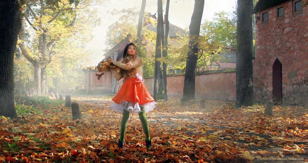 Обои для рабочего стола Светловолосая девочка, стоящая посреди городской улицы, усыпанной осенними листьями, держащая в руках охапку таких же листьев, автор Алексей Макаренок