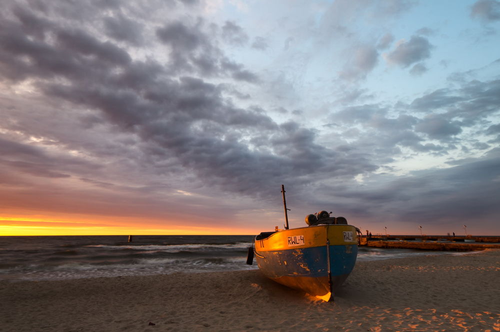 Обои для рабочего стола Рыбацкий баркас, стоящий на песчаном, морском побережье, освещенный лучами заходящего за линию горизонта солнца на вечернем небосклоне с серыми, перистыми облаками, автор Mariuszbrcz
