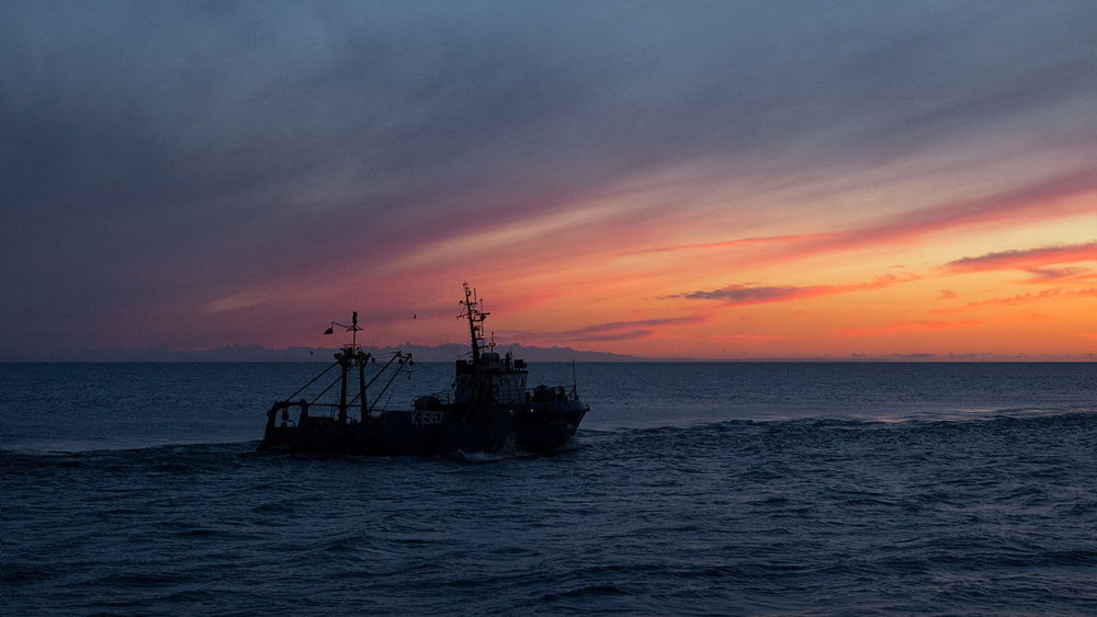 Обои для рабочего стола Рыбацкий баркас, плывущий по морю в окружении парящих в воздухе чаек на фоне заката на вечернем небосклоне, baltiec