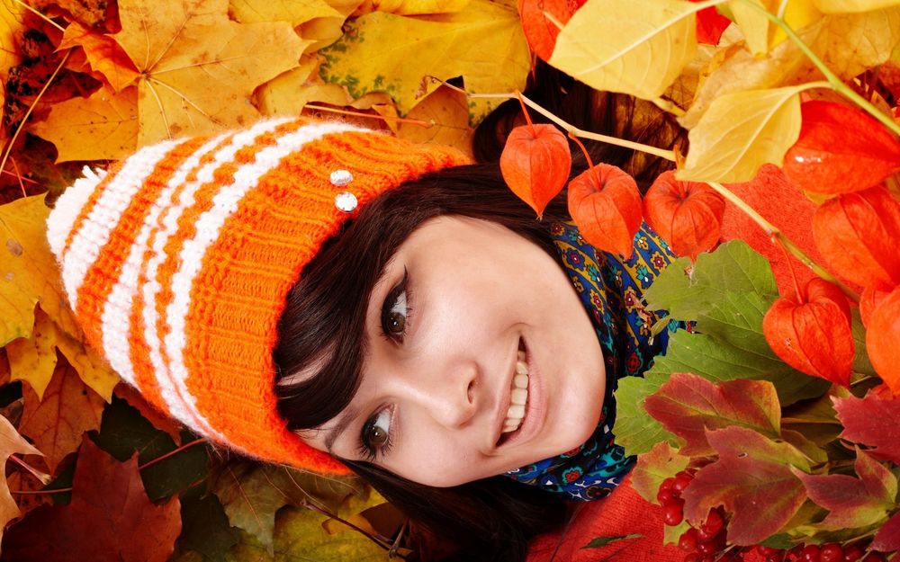 Обои для рабочего стола Улыбающаяся девушка в яркой шапочке лежит в осенних листьях