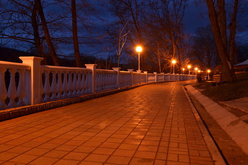 Обои на рабочий стол Мостовая, покрытая каменной плиткой с фигурным ограждением, ярко освещенная уличными фонарями, растущими по краям деревьями на фоне ночного, пасмурного неба