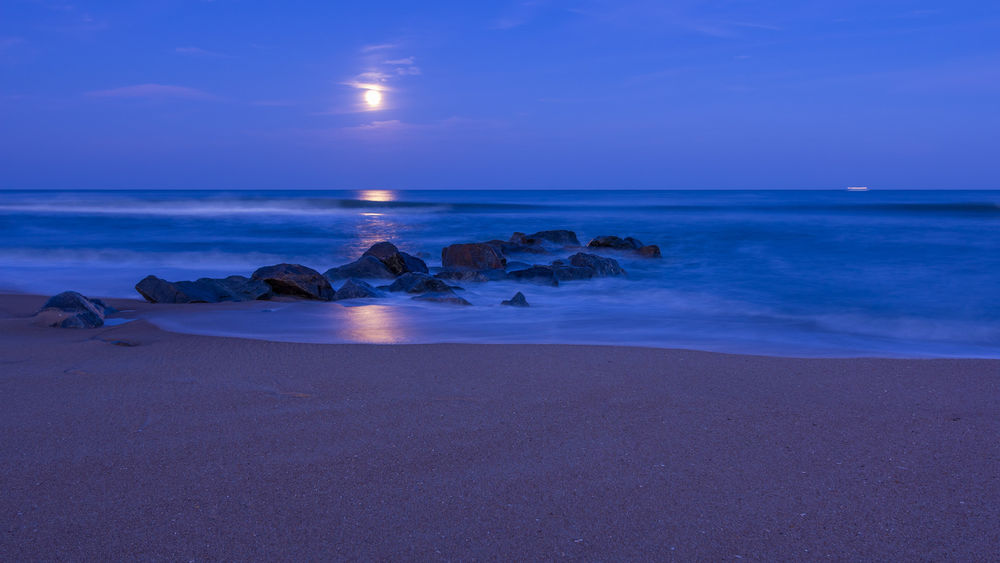 Обои для рабочего стола Ночной пляж с камнями в воде моря, над которым светит полная Луна, отражаясь в воде