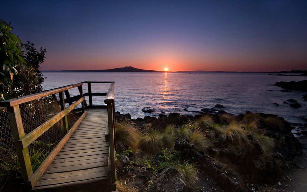 Обои для рабочего стола Деревянный мостик, стоящий на берегу озера на фоне горного образования на линии горизонта и заходящего солнца на вечернем небосклоне