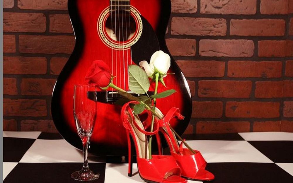 Обои для рабочего стола Красные туфли, гитара, бокал и розы стоят на столе в клеточку, на фоне кирпичной стены