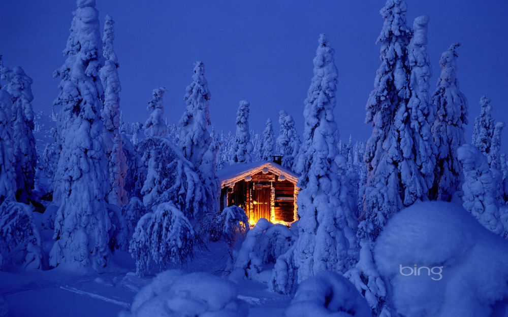 Обои для рабочего стола Деревянный, охотничий домик, ярко освещенный электрическим светом, засыпанный густым слоем снега в окружении елей (bing)