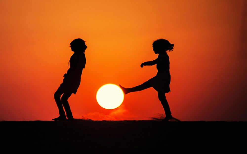 Обои для рабочего стола Силуэты детей на фоне заходящего солнца, девочка приложила ногу к солнцу, имитируя удар по мячу