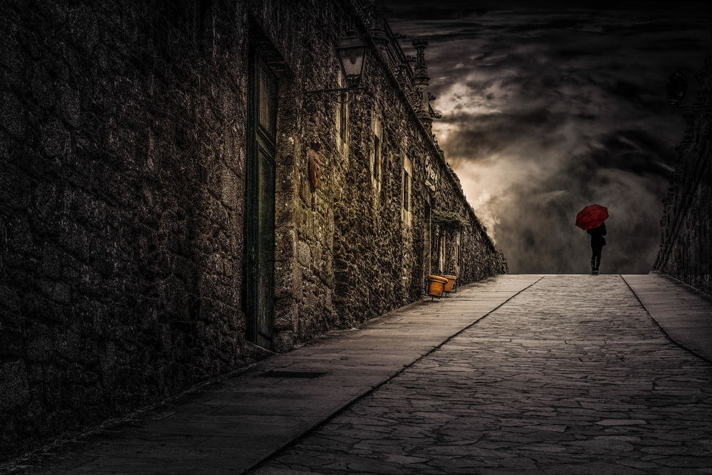 Обои для рабочего стола Стройная девушка в темной одежде, держащая в руке красный зонтик, идущая по мощеной мостовой среди мрачных стен зданий на фоне вечернего небосклона с темными, дождевыми облаками