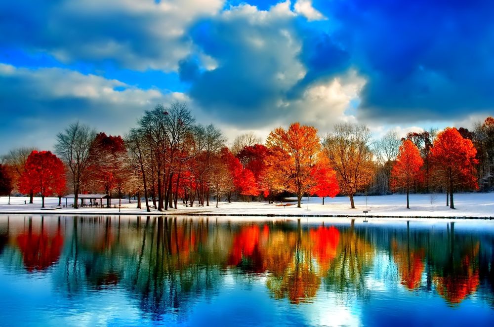 Обои для рабочего стола Деревья с красной, осенней листвой, отразившиеся в зеркальной глади водоема, растущие на берегу, покрытом свежевыпавшим снегом на фоне неба с разноцветными облаками