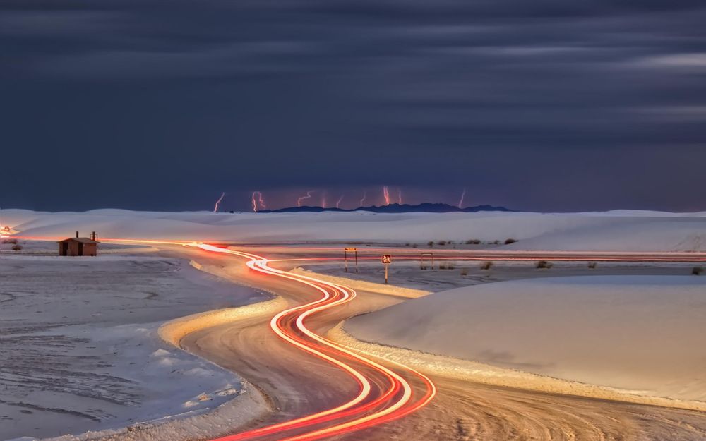 Обои для рабочего стола Извилистая дорога со световыми полосками, проходящая через заснеженное поле на фоне ночного, грозового неба со сверкающими молниями на линии горизонта