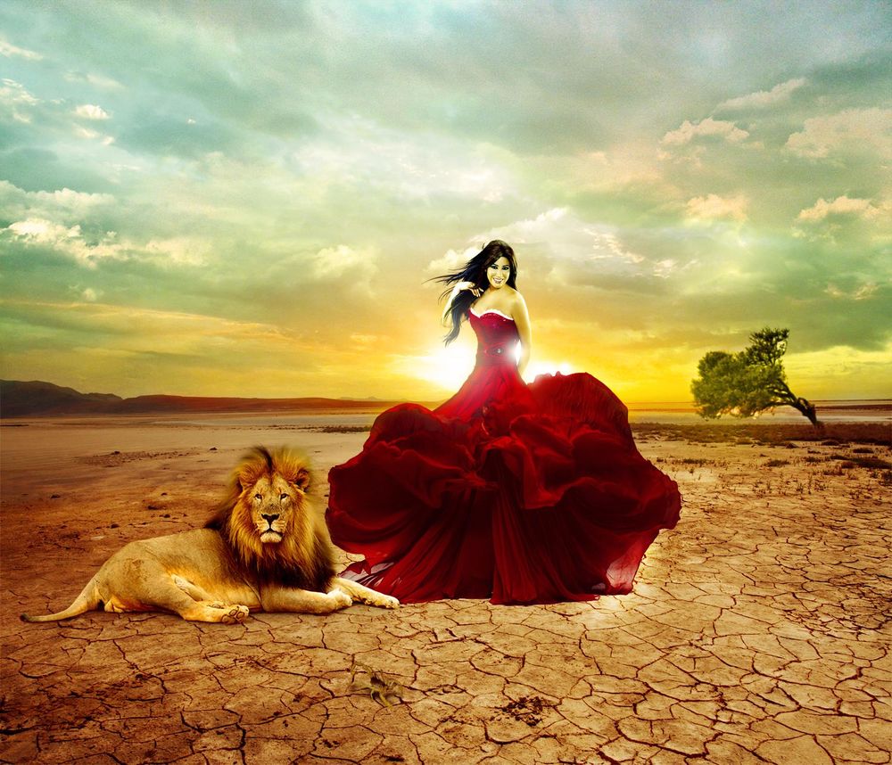 Обои для рабочего стола Девушка в пышном длинном красном платье, стоит улыбаясь на фоне неба с заходящим Солнцем, на потрескавшейся от жары земле рядом с лежащим львом