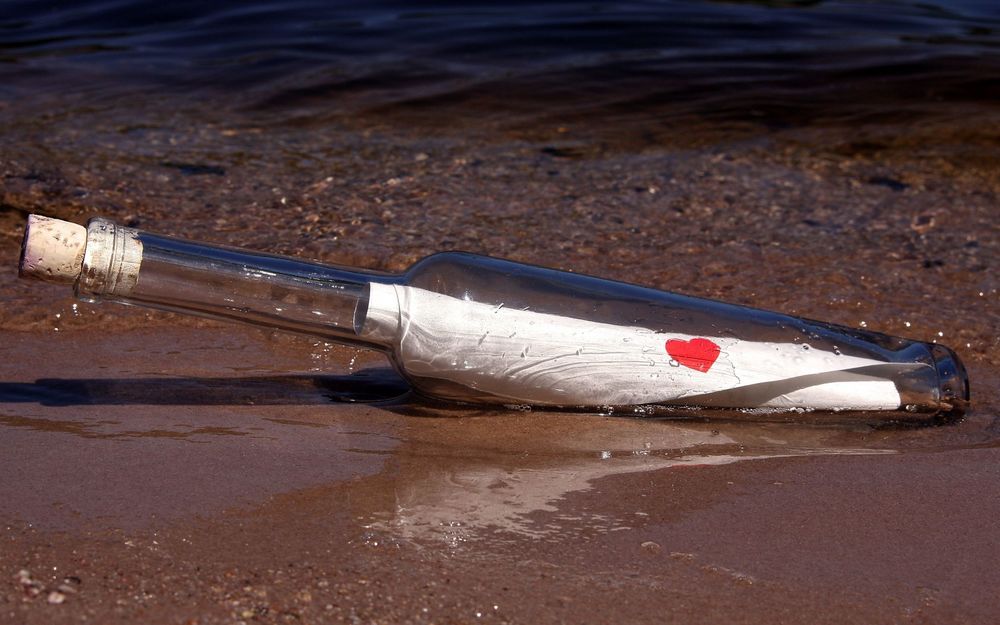Обои для рабочего стола Стеклянная бутылка с длинным, узким горлышком, закрытая пробкой с лежащим в ней листком белой, свернутой бумаги с нарисованным красным сердечком, лежащая на песчаном берегу моря