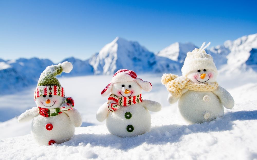 Обои для рабочего стола Три игрушечных снеговичка стоят на снегу на фоне горы и неба