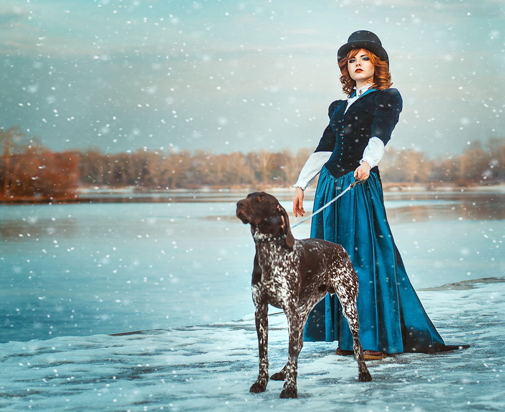 Обои для рабочего стола Рыжеволосая девушка в черной шляпе, держащая на поводке породистую собаку, стоящая на берегу озера под падающим снегом, автор Галия Желнова