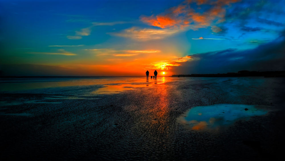 Обои для рабочего стола Силуэты двух людей, совершающих прогулку со своими собаками по мелководью морского залива на фоне заката солнца на вечернем небосклоне с разноцветными облаками