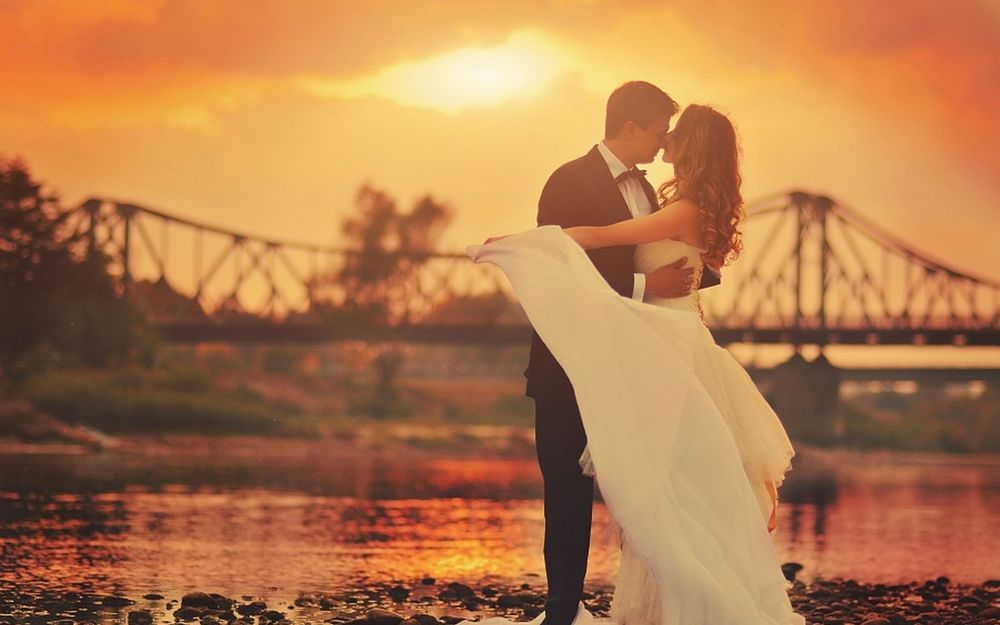 Обои для рабочего стола Мужчина и девушка обнявшись стоят у реки на закате солнца