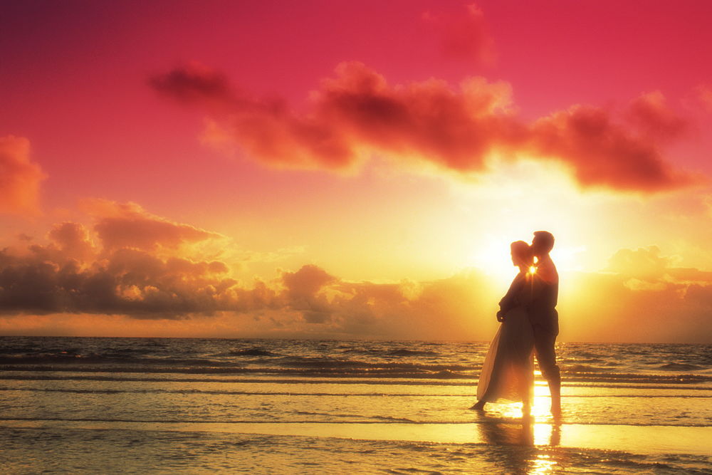 Обои для рабочего стола Мужчина и девушка стоят обнявшись, на берегу в прибрежных волнах, на фоне заката