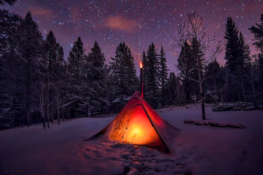 Обои для рабочего стола В заснеженном лесу под ночным звездным небом стоит палатка, внутри освещенная светом, на верхушке палатки горит факел, автор Lars Laber