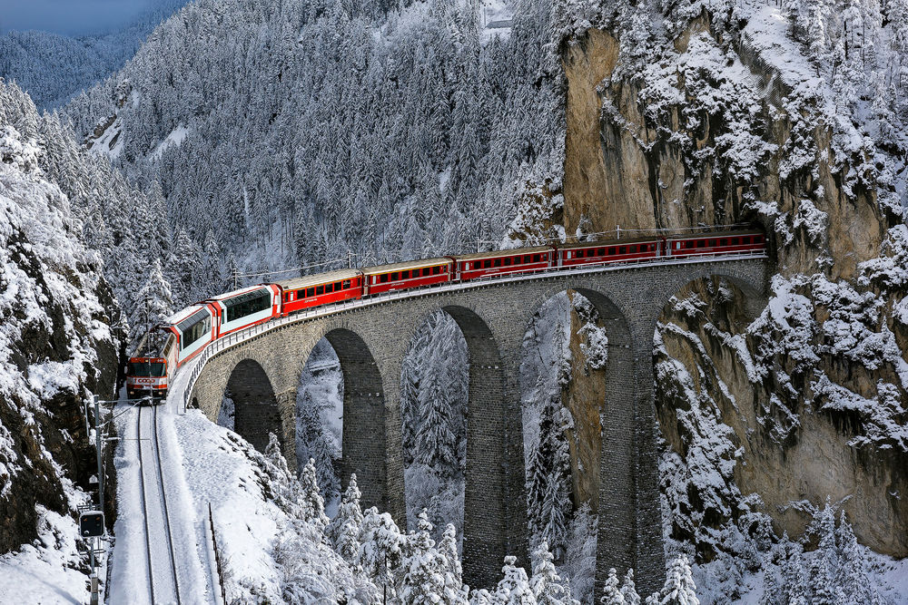 Обои для рабочего стола Пассажирский поезд, выходящий из туннеля в горах, идущий по железнодорожному мосту, проложенному через заснеженное, горное ущелье