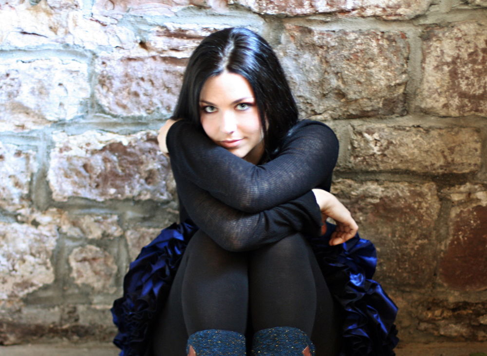 Обои для рабочего стола Эми Линн Харцлер / Amy Lynn Hartzler/, американская певица-поэтесса и пианистка, вокалистка группы Evanescence сидит возле каменной стены, сложив руки на коленях