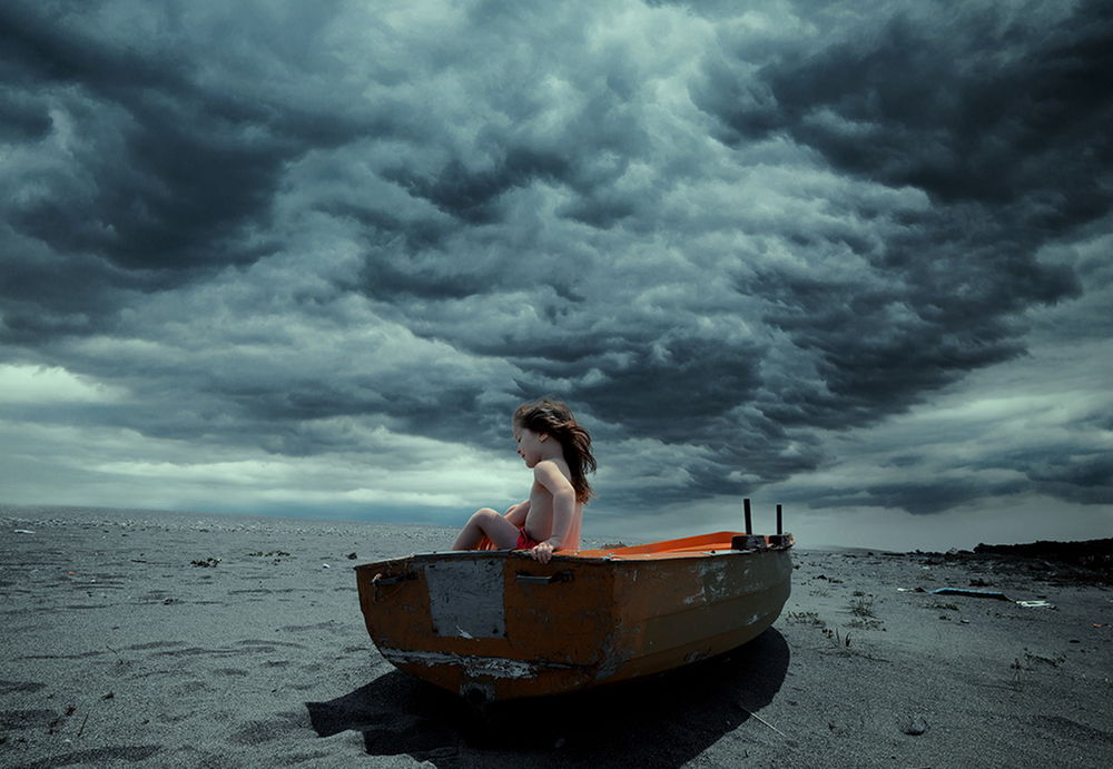 Обои для рабочего стола Девочка, сидящая на борту лодки, стоящей на песке на фоне пасмурного небосклона с темно-серыми облаками, автор Garas Ionut