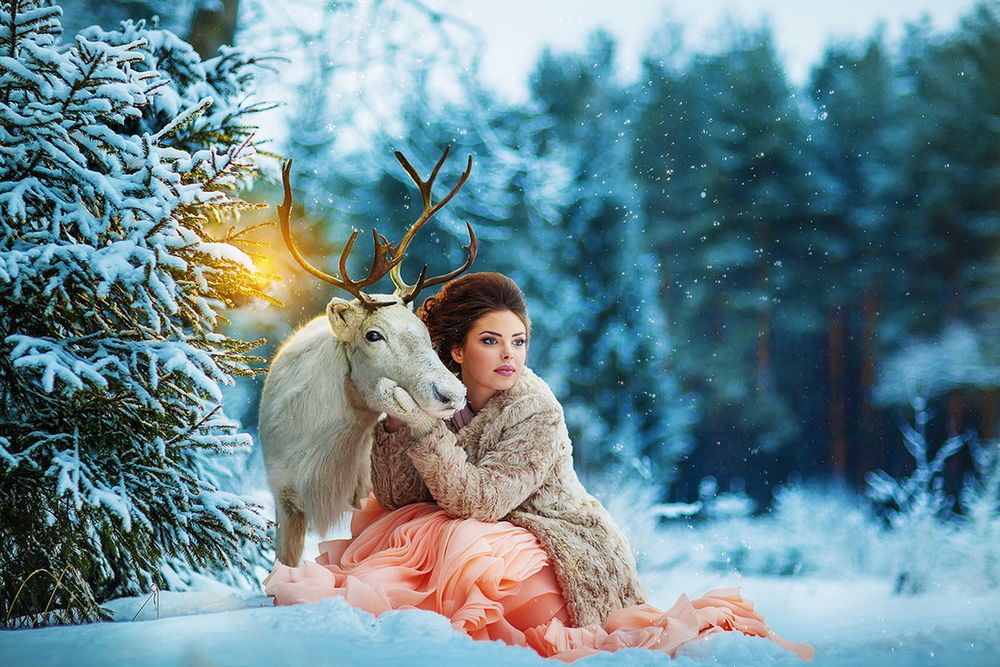 Обои для рабочего стола Красивая девушка, держащая за голову белого оленя с ветвистыми рогами, находящаяся на заснеженной, лесной опушке, автор Илья Двояковский