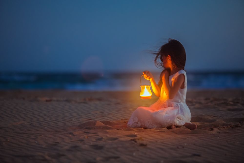 Обои для рабочего стола Темноволосая девочка в белом платье, держащая в руке фонарь с ярко горящим фитилем, сидящая на песчаном берегу моря на фоне вечернего небосклона