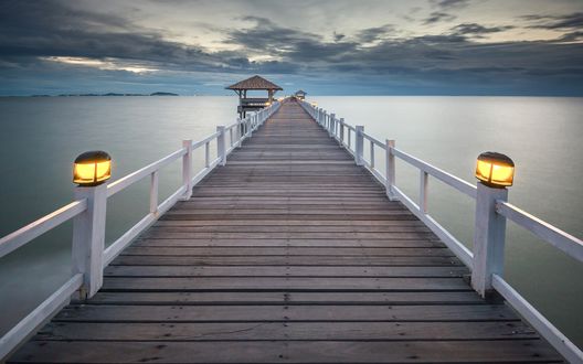 Обои на рабочий стол Деревянный мост с горящими на его перилах фонарями, уходящий от берега морского залива на фоне вечернего небосклона с темными облаками