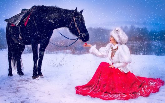 Обои на рабочий стол Девушка в белой, меховой шапке и жилетке, пышной, красной юбке, сидящая на корточках на снегу перед стоящим возле нее статным, вороным конем на фоне падающего снега