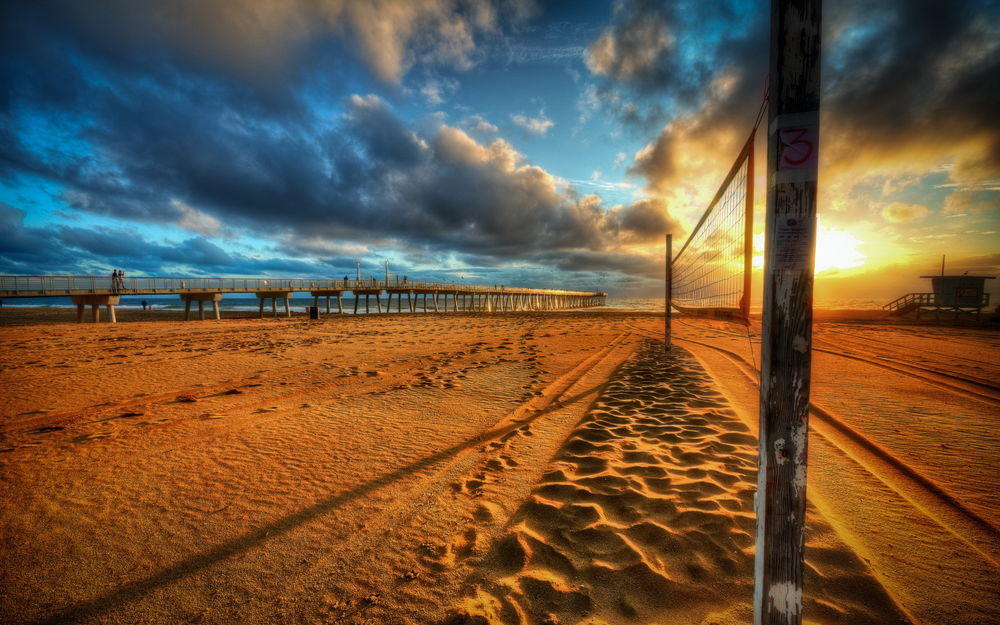 Обои для рабочего стола Песчаный пляж с натянутой между двумя деревянными столбами сеткой для пляжного волейбола, на берегу морского залива на рассвете, каменным мостом, уходящим в море