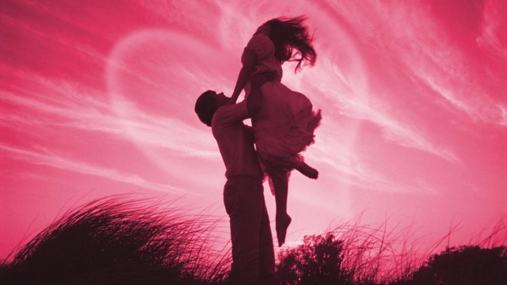 Обои для рабочего стола Влюбленный мужчина поднял на руки любимую женщину на фоне розового неба с огромным сердцем