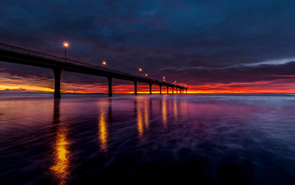 Обои для рабочего стола Мост с горящими на нем уличными фонарями, проходящий через морской залив на фоне багряной, закатной полоски на вечернем небосклоне с темными, грозовыми облаками