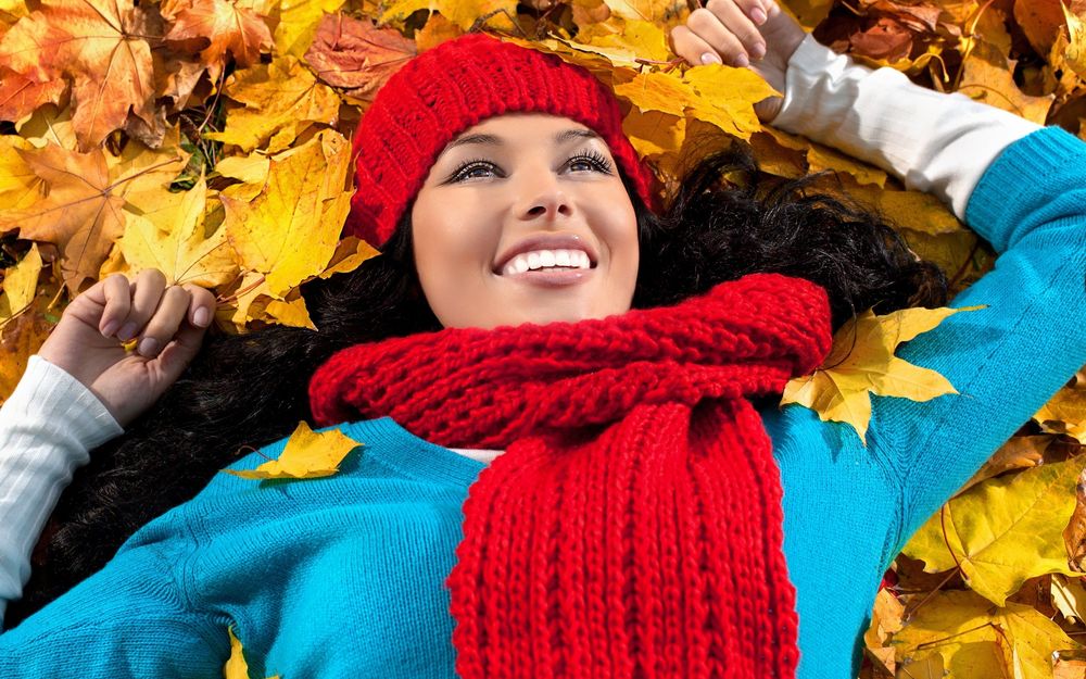 Обои для рабочего стола Улыбающаяся девушка в красной шапочке и шарфике лежит на осенних листьях