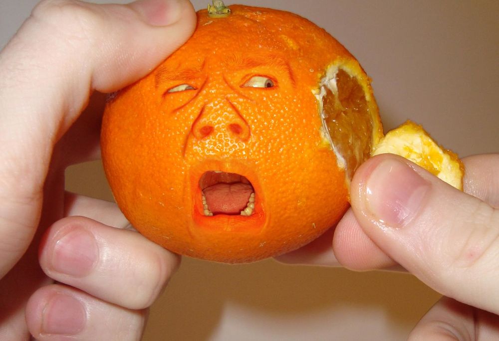 Обои для рабочего стола Человеческие руки снимают кожуру с апельсина, у которого глаза, нос и рот, кричащий от возмущения и боли