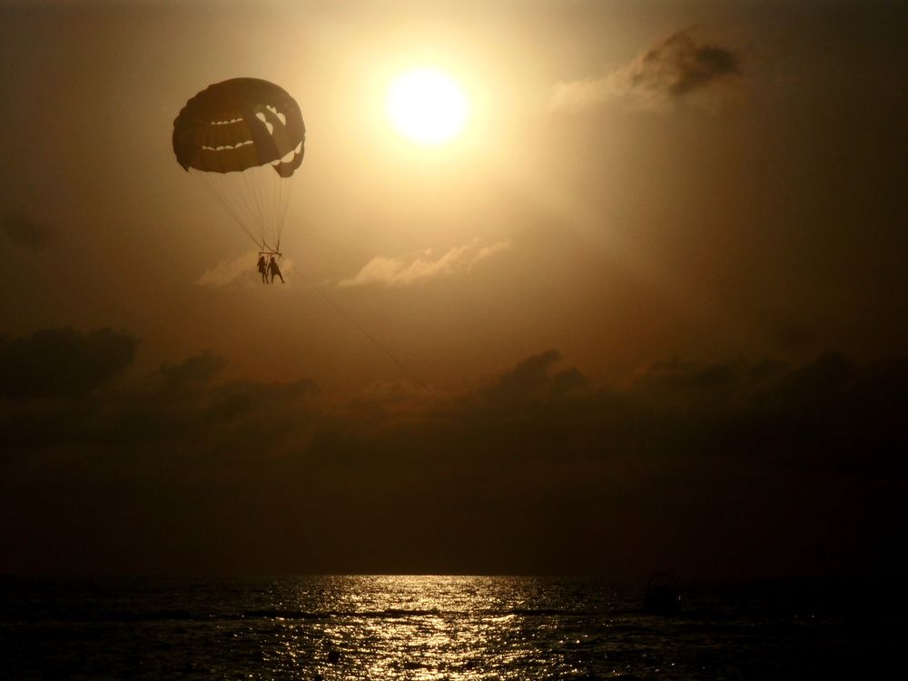 Обои для рабочего стола Влюбленные парень и девушка, парящие в воздухе под куполом парашюта над озером на фоне ослепительного, солнечного диска на вечернем небосклоне