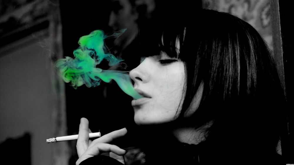 Обои для рабочего стола Девушка курит, держа в руке сигарету и выпуская со рта зеленый дым