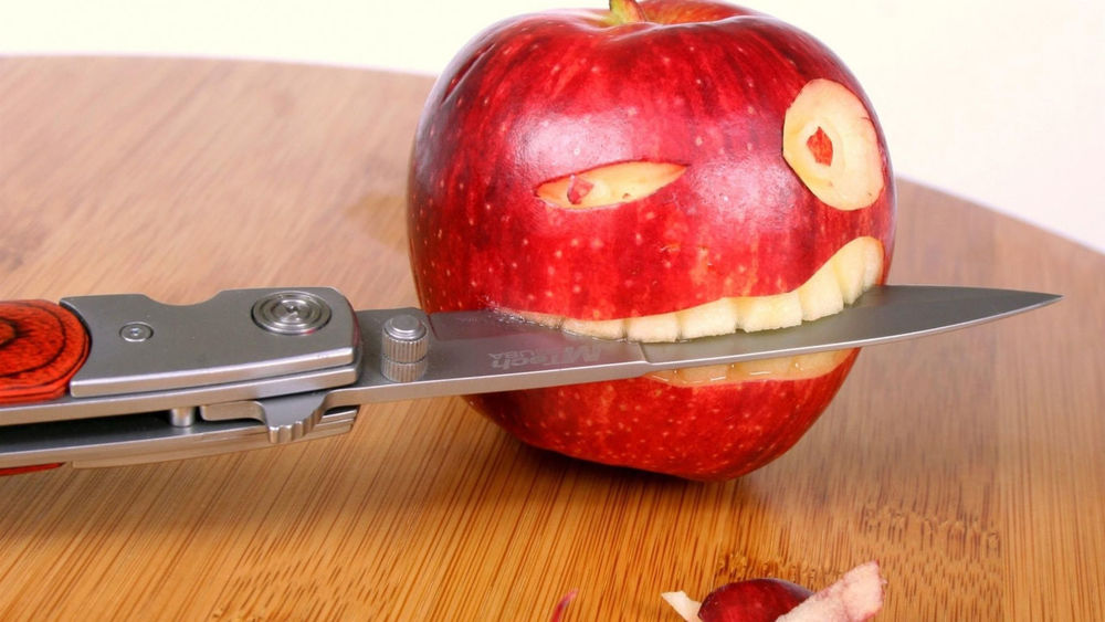 Обои для рабочего стола Красное яблоко с вырезанными глазами и ртом, в котором вставлено лезвие складного ножа