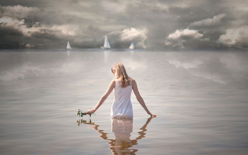 Обои для рабочего стола Девушка в белом платье стоящая в воде, держит в руке букетик цветов, вдалеке виднеются парусники