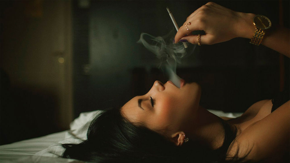 Обои для рабочего стола Девушка лежа на спине, держит в руке зажженную сигарету и выпускает дым со рта