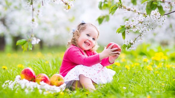 Обои на рабочий стол Радостная девочка сидит на цветочной полянке с яблоком в руках, среди цветущего сада